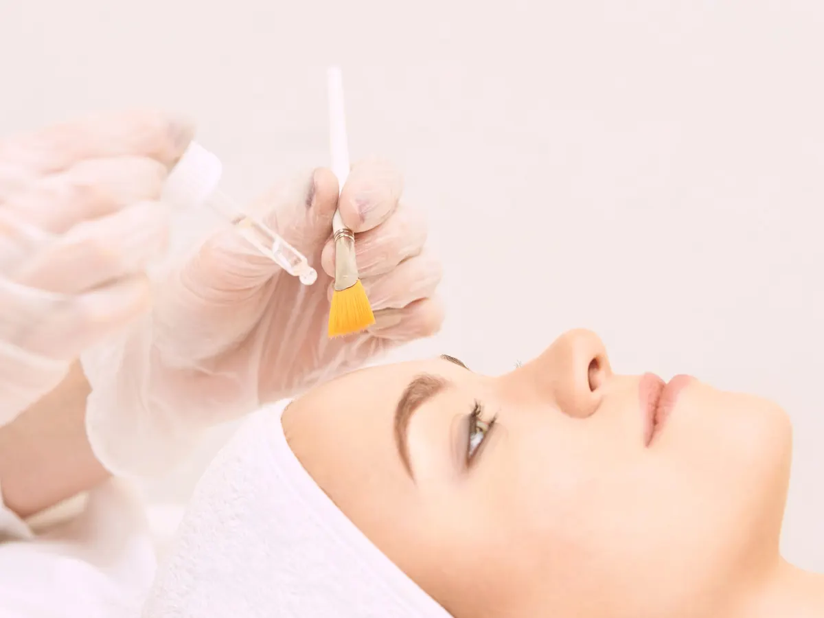 Stock photograph of a model: a woman lying back in a white head wrap as gloved hands hold a dropper and a brush to apply a peel