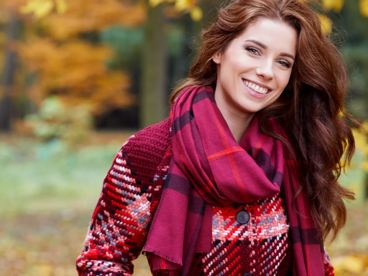Stock photograph of a model: a smiling woman with long auburn hair in a plaid scarf and knit coat stands among autumn trees