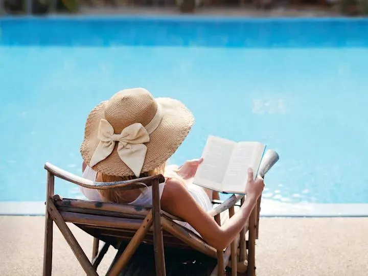 Stock photograph of a model: a woman in a sun hat reads in a lounge chair beside a swimming pool