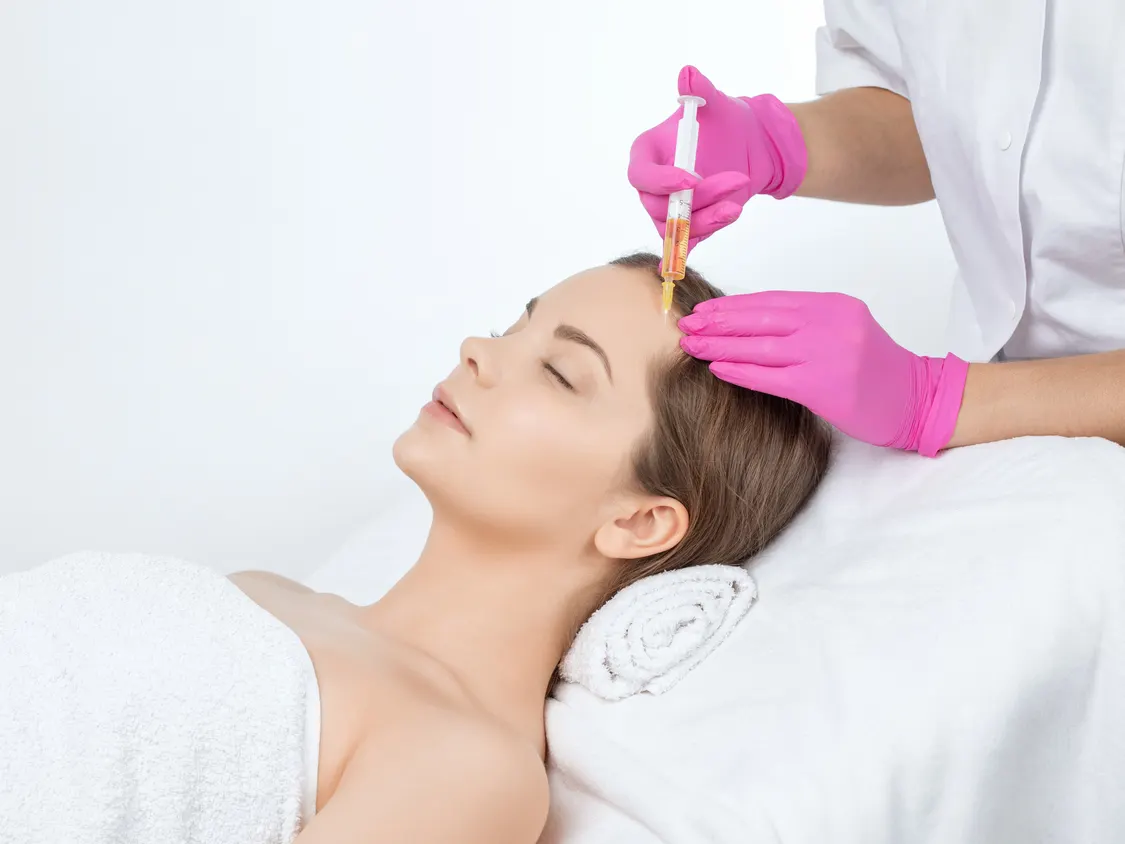 Stock photograph of models: a woman lying on a treatment bed receives an injection in her forehead from a clinician in pink gloves