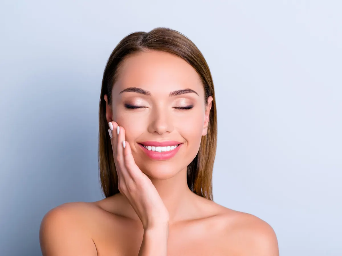 Stock photograph of a model: a smiling woman with her eyes closed rests a hand on her cheek against a pale blue background
