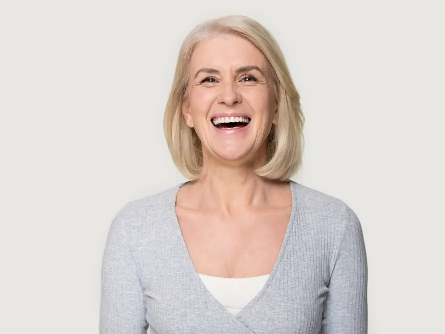 Stock photograph of a model: a laughing older woman with a blonde bob and a grey cardigan against a plain backdrop