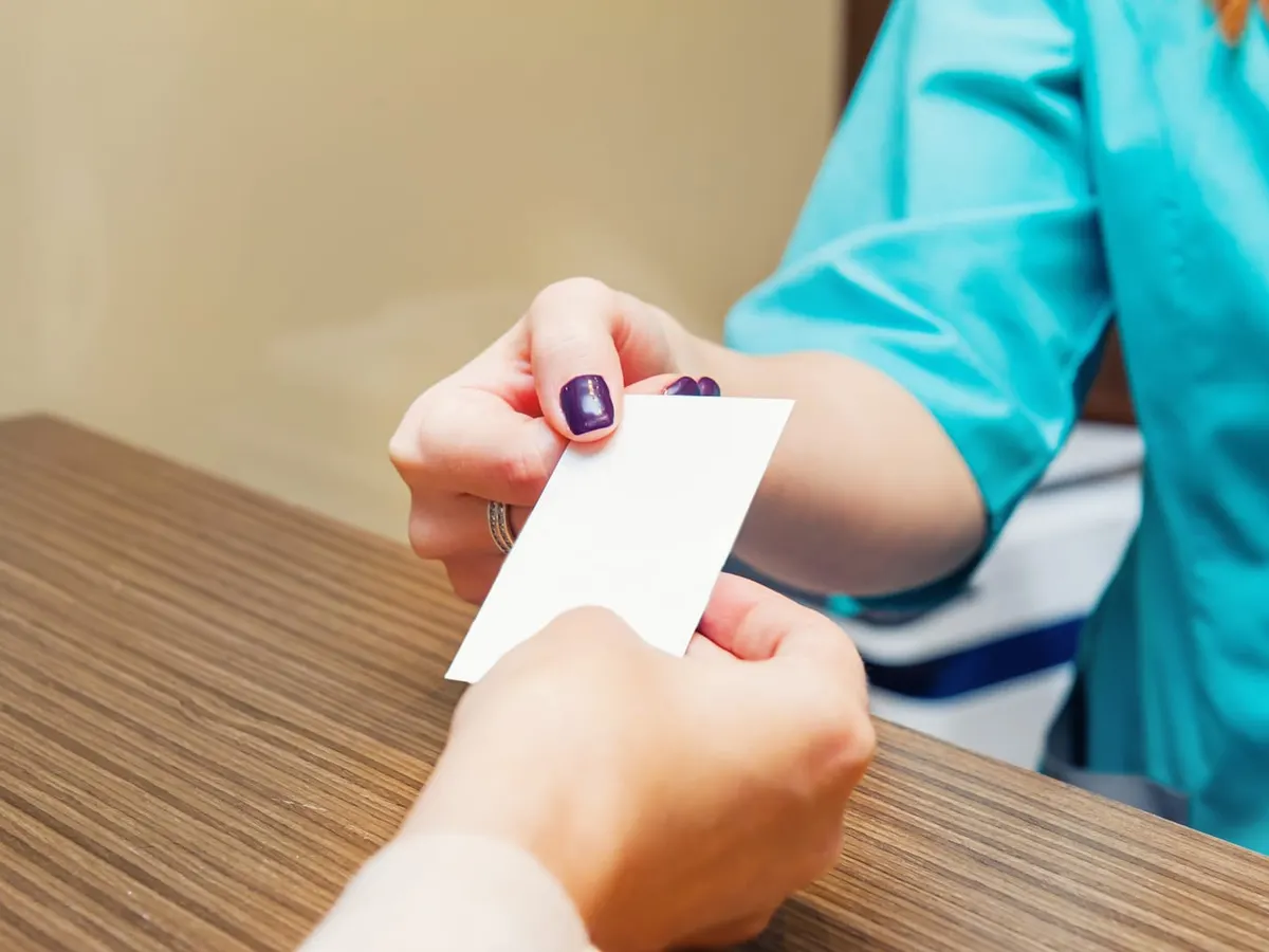 Stock photograph of a model: a clinic receptionist in teal scrubs hands a blank card across a wooden front desk to a patient.