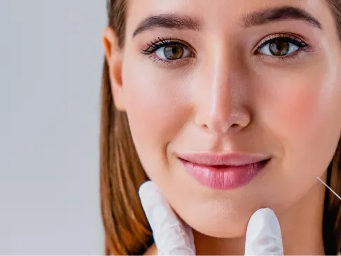Stock photograph of a model: a woman with straight brown hair smiles while gloved hands hold her chin and a syringe waits beside her