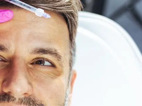 Stock photograph of a model: a pink-gloved hand holds a syringe above the forehead of a bearded man in a treatment chair