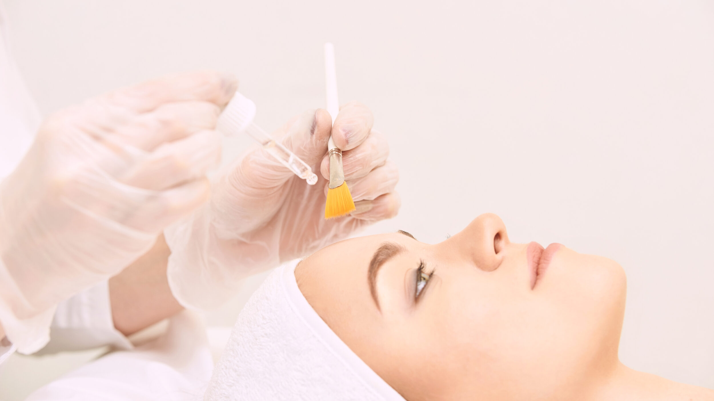 Stock photograph of a model: a woman lying back in a white head wrap as gloved hands hold a dropper and a brush to apply a peel