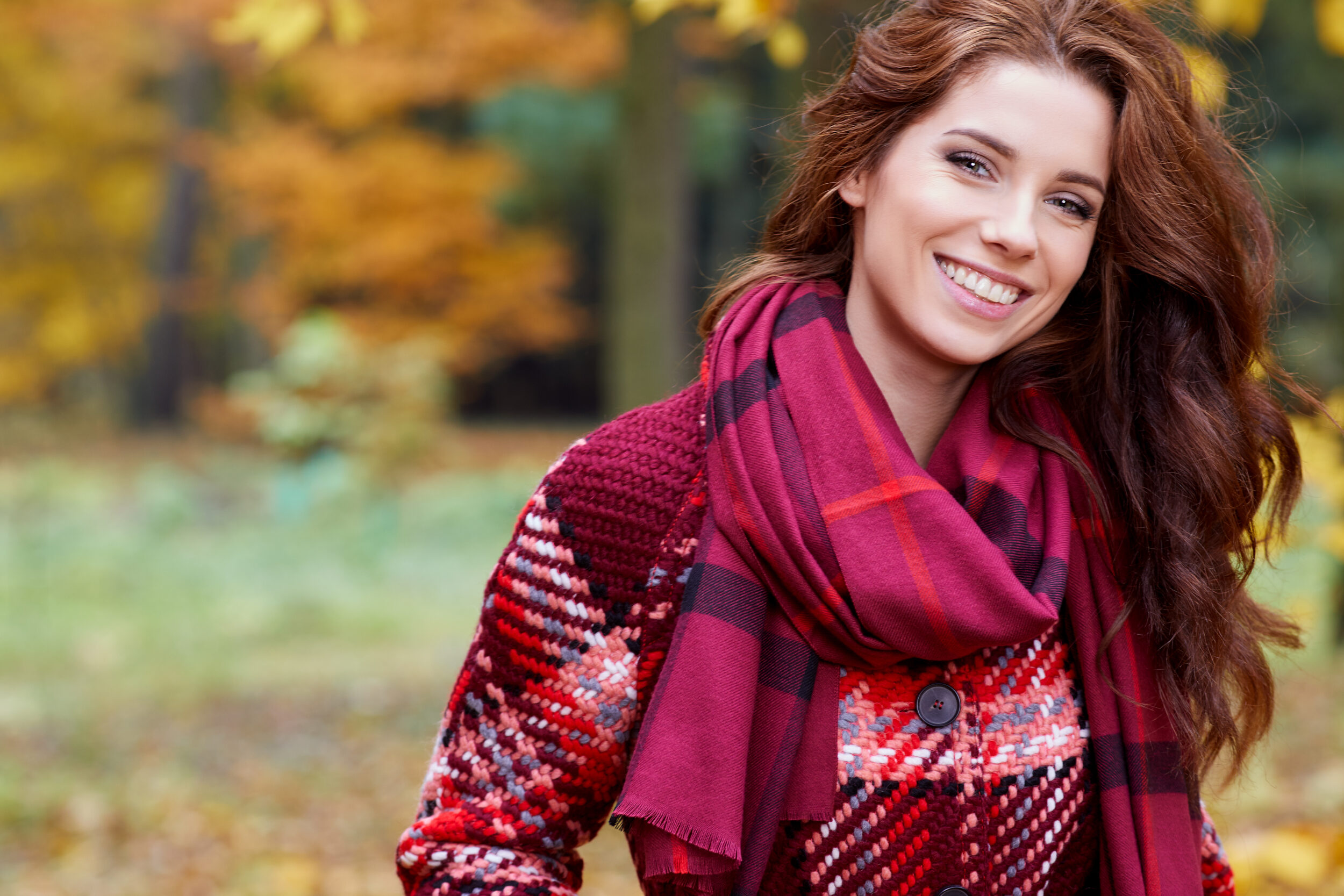 Stock photograph of a model: a smiling woman with long auburn hair in a plaid scarf and knit coat stands among autumn trees