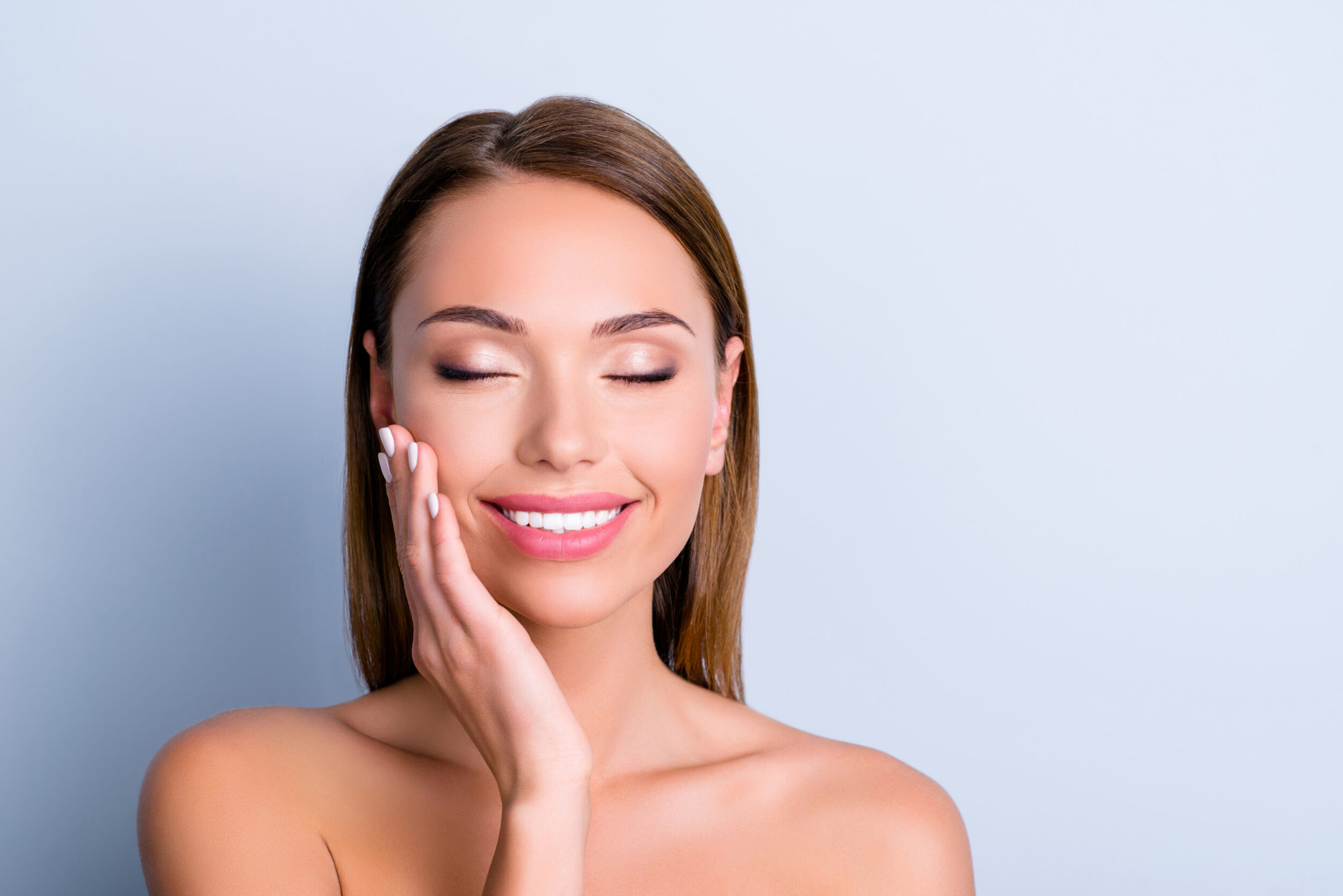 Stock photograph of a model: a smiling woman with her eyes closed rests a hand on her cheek against a pale blue background
