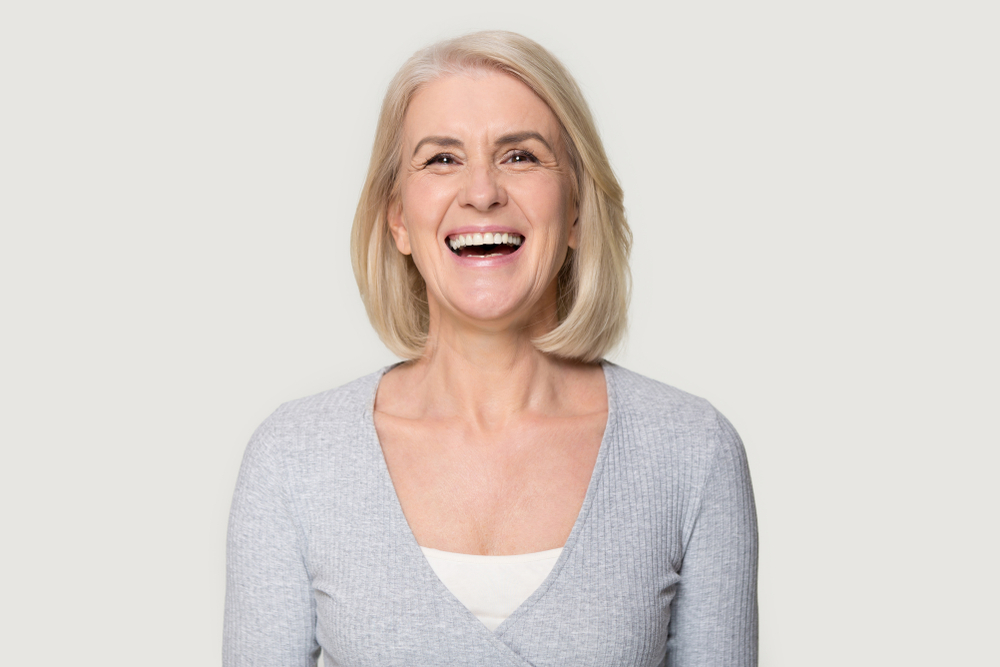 Stock photograph of a model: a laughing older woman with a blonde bob and a grey cardigan against a plain backdrop