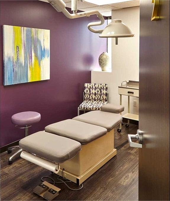 An exam room at Dermatology Specialists of Dallas: a treatment chair under a surgical light, beside a plum wall hung with an abstract painting
