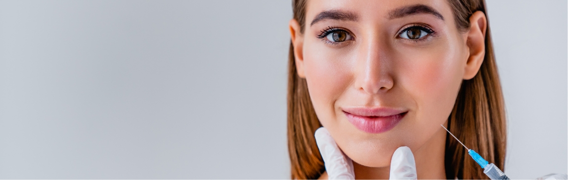 Stock photograph of a model: a woman with straight brown hair smiles while gloved hands hold her chin and a syringe waits beside her