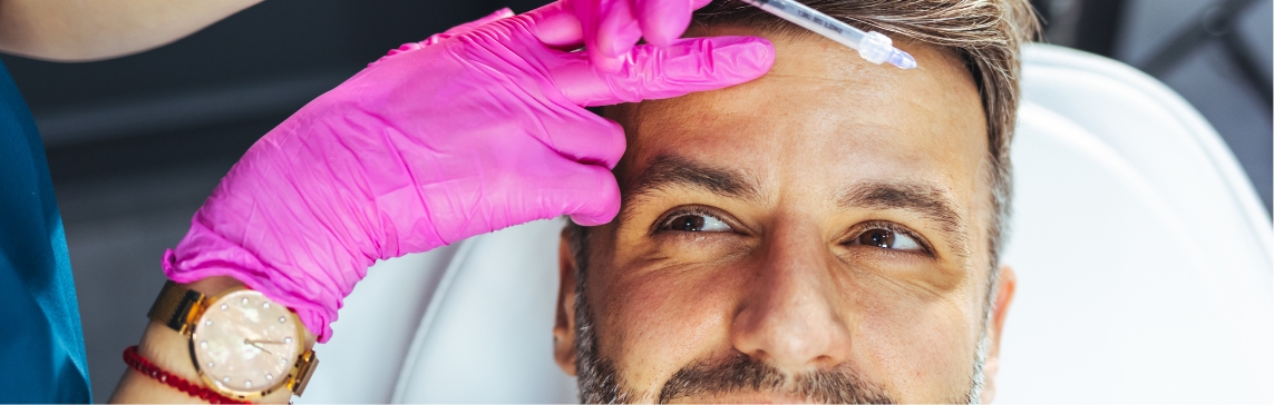 Stock photograph of a model: a pink-gloved hand holds a syringe above the forehead of a bearded man in a treatment chair