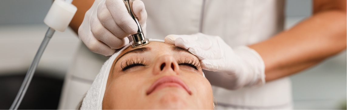 Stock photograph of a model: a woman with her eyes closed and a head towel receives microdermabrasion with a metal handpiece on her forehead